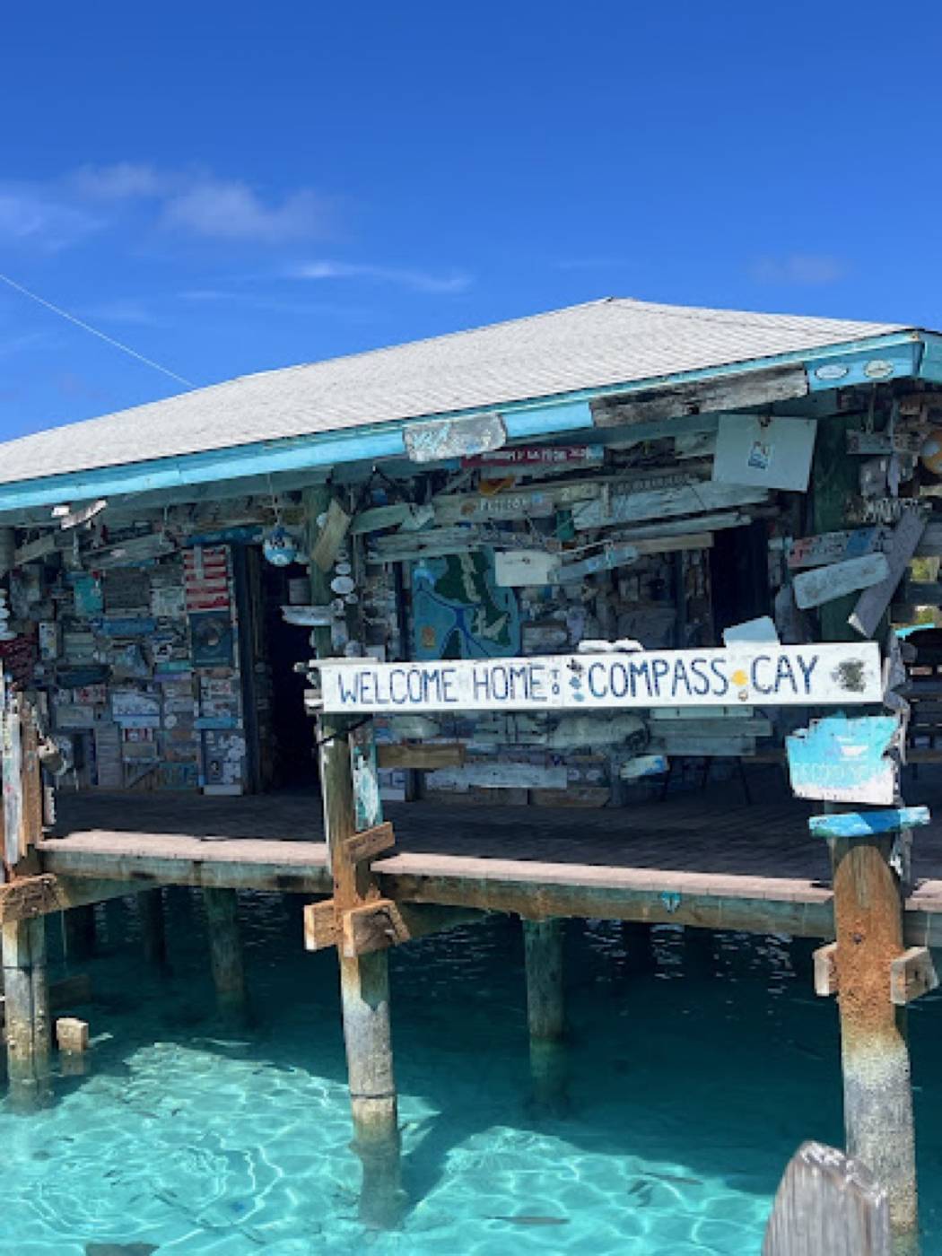 Wooden dock house with a 'Welcome Home Compass Cay' sign above clear water