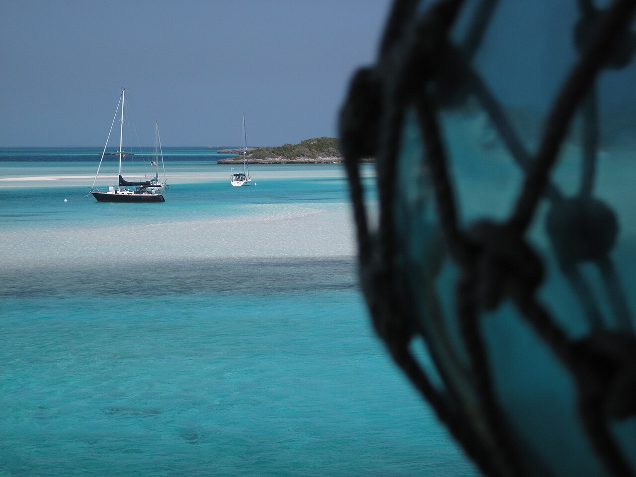 Two sailboats at anchor in pale turquoise water near Warderick Wells, seen past a glass float