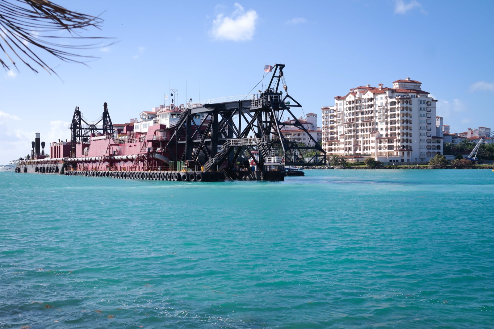A dredge with a crane working at the entrance to Government Cut, Miami, with turquoise water in front and waterfront towers behind