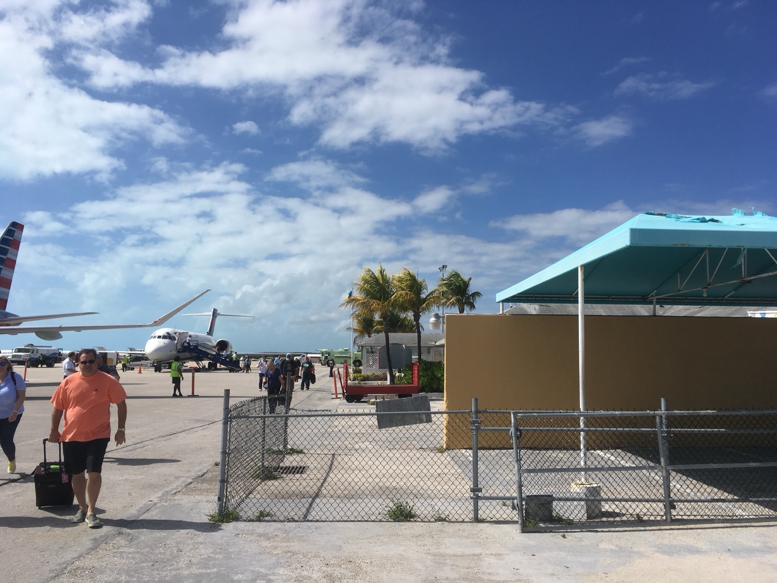 Passengers walk across the ramp at Exuma International Airport beside a parked regional jet, with a palm-lined terminal building on the right