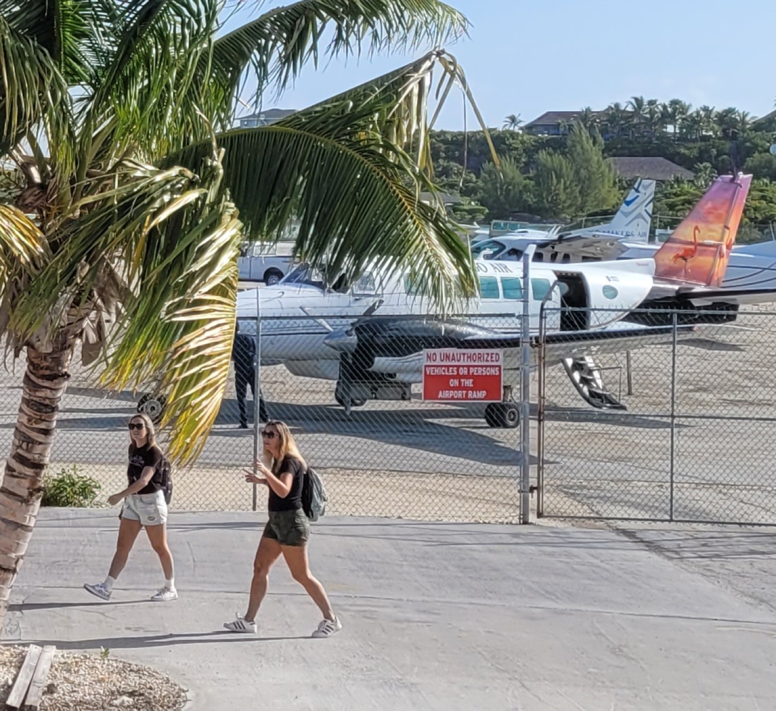 Two women walk past a no-unauthorized-persons sign on the Staniel Cay Airport apron, with small Flamingo Air aircraft parked behind a fence