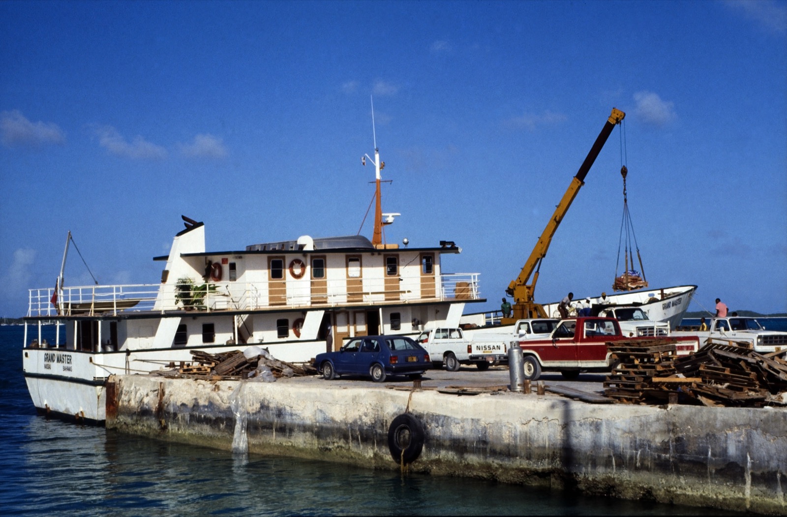 The mail boat Grand Master tied up at a quay with pickup trucks and a crane lifting cargo, in a 1989 photograph