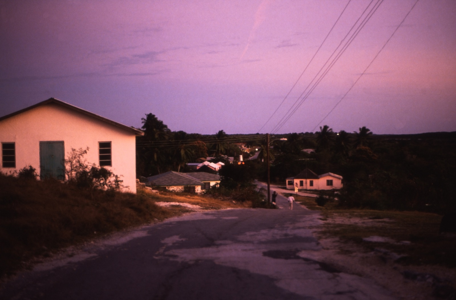 Dusk view of a paved village road in George Town, Great Exuma, with small white houses and a few distant people