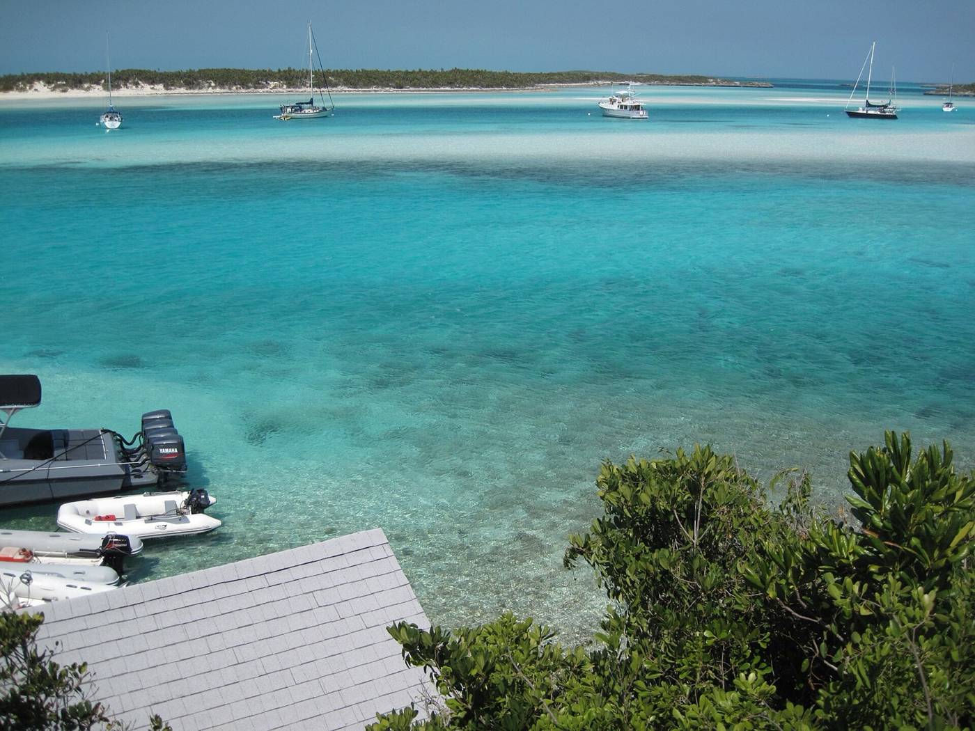 Sailboats and a motor yacht anchored in turquoise water off a sandy Exuma shore