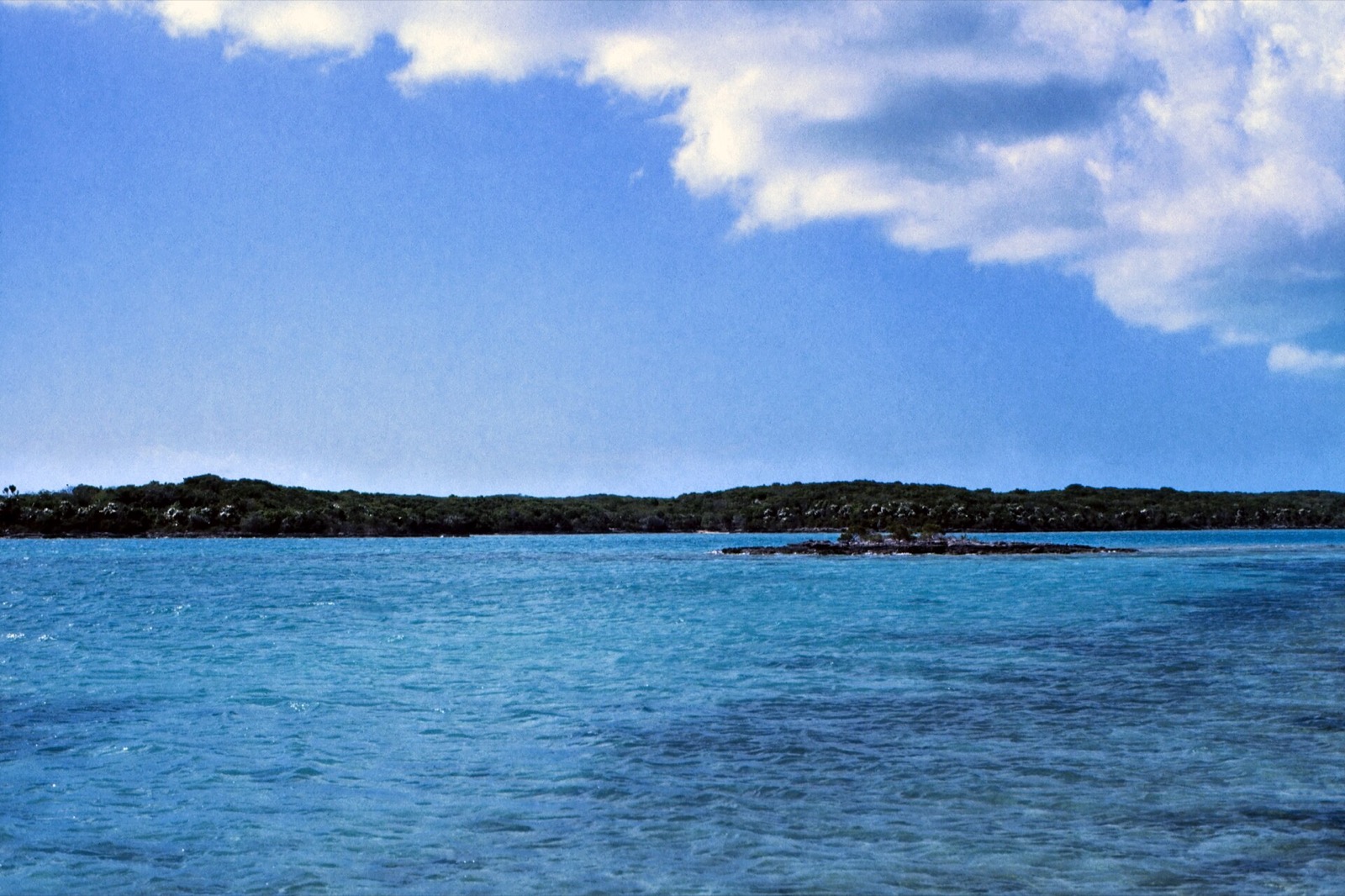 Low wooded shoreline and a small rocky cay across turquoise water off Great Exuma's northeast coast