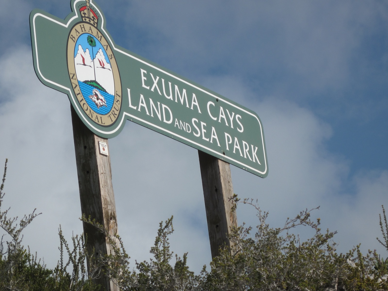 Green and white Exuma Cays Land and Sea Park sign with the Bahamas National Trust crest, on wooden posts against a cloudy sky