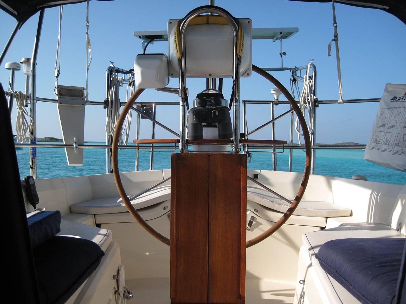 Sailboat helm with a steering wheel, binoculars and a VHF handset over turquoise Bahamas water