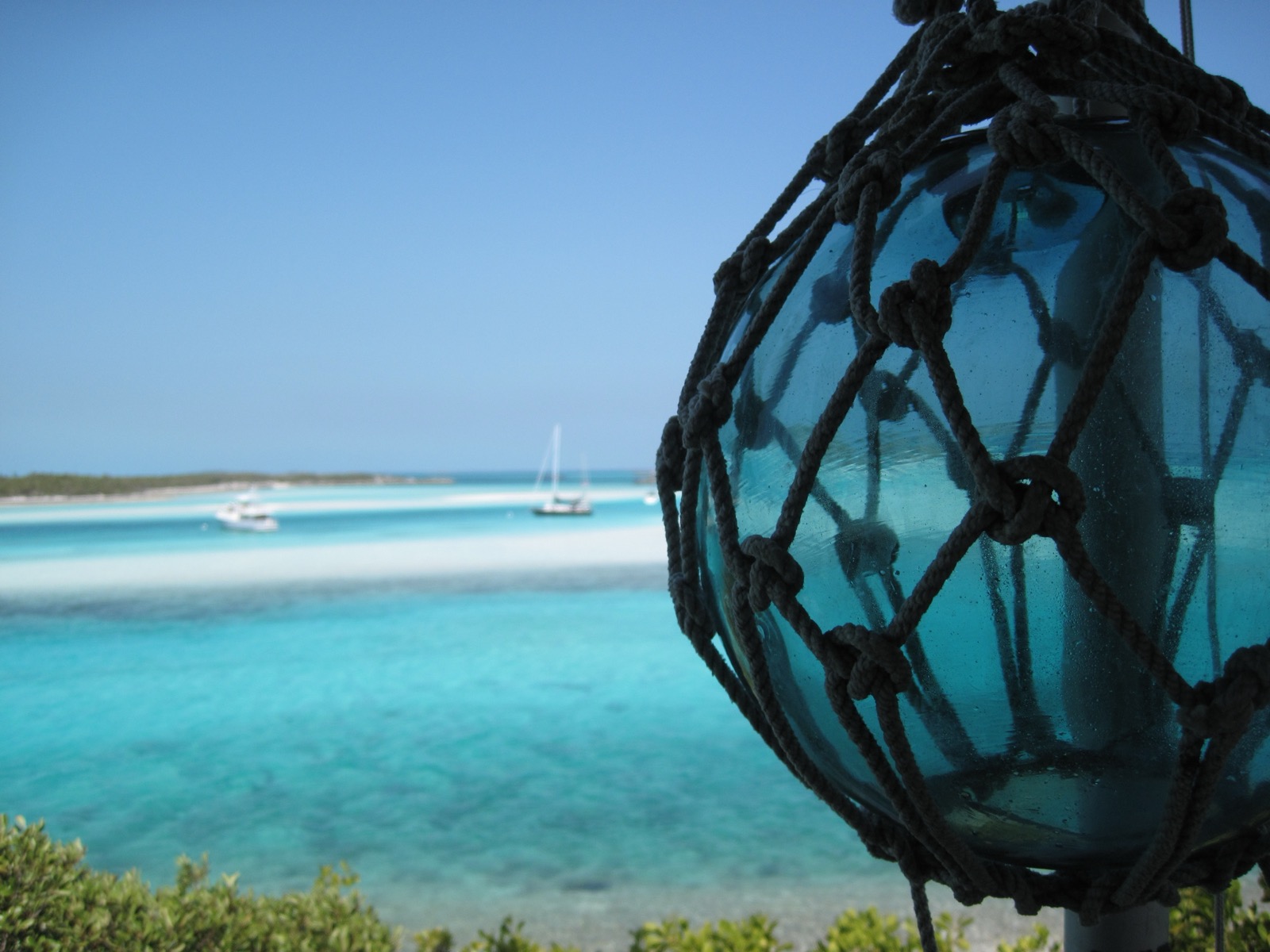 View from Warderick Wells across turquoise water and a white sandbar to sailboats, with a glass float in the foreground